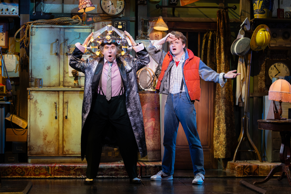 Two actors performing in a theatrical setting, one wearing a sparkly coat with lights on their head, and the other in a red vest and distressed denim, both appear surprised.