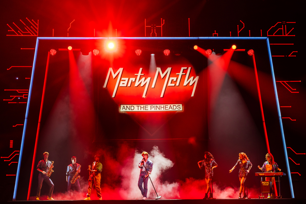 A vibrant stage performance featuring a band named 'Marty McFly and the Pinheads,' with musicians playing instruments and singers performing under dramatic lighting.