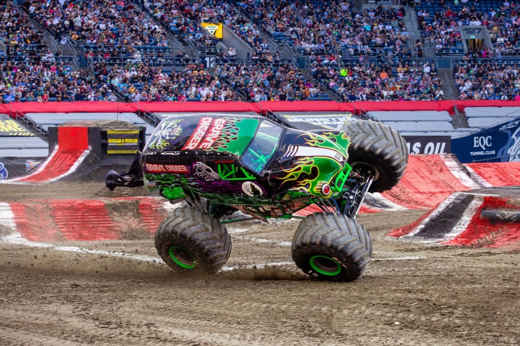 A monster truck performing a wheelie on a dirt track during a stunt event, with a large crowd in the background.