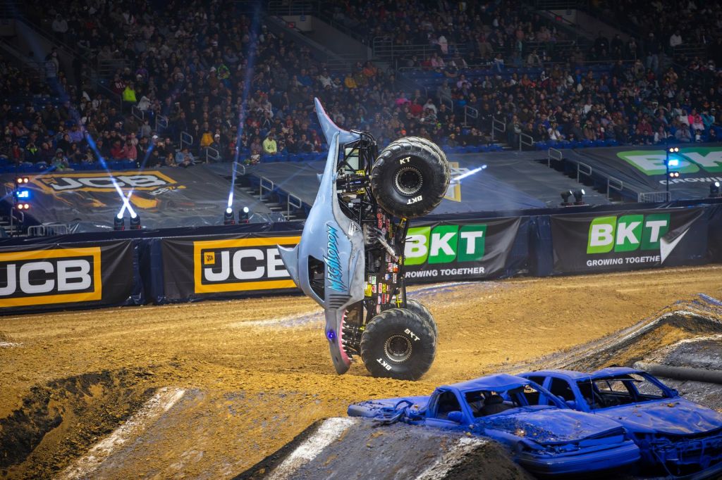 A monster truck performing a wheelie at a live motorsport event, with an audience in the background and crushed cars in the foreground.