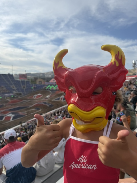 A child wearing a bright red bull mask with yellow accents, giving two thumbs up while sitting in a stadium filled with spectators at a motorsport event.