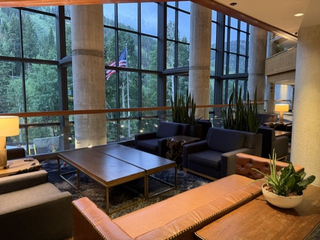 Modern lounge area with large windows showcasing a forest view, featuring comfortable seating, a coffee table, and decorative plants.