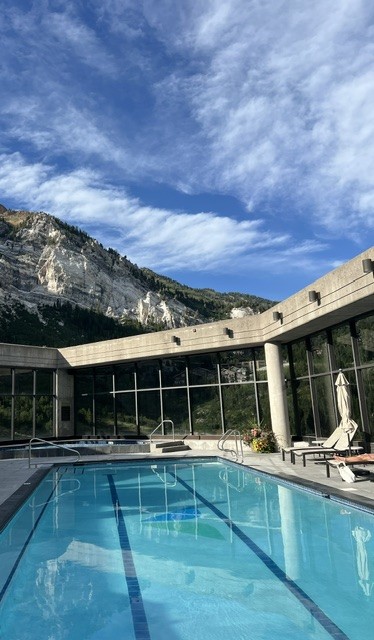 Indoor swimming pool with large windows, surrounded by mountains and blue sky.