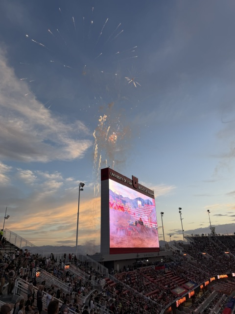 Fireworks display over a large video screen at a stadium during sunset, with spectators visible in the stands.