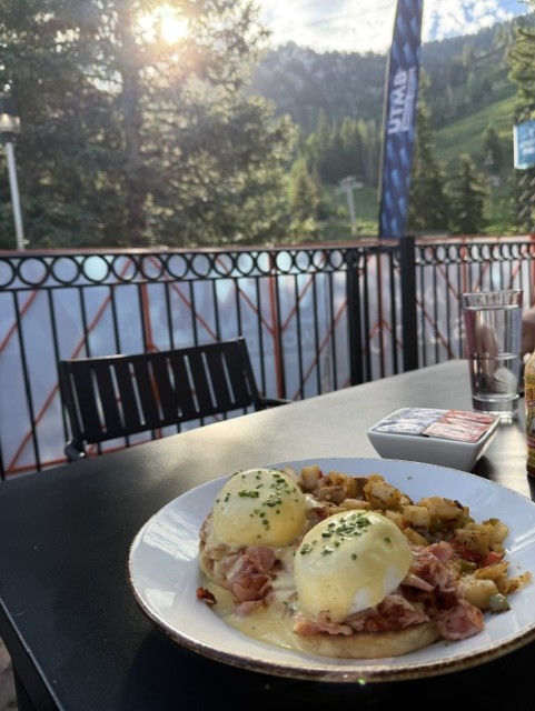 Plate of eggs benedict with ham and hollandaise sauce, served with a side of hash browns, set on an outdoor table with a scenic mountain view.