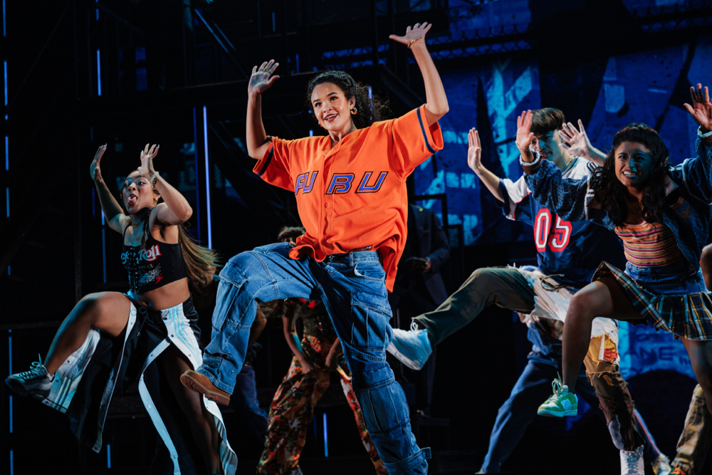 A group of dancers performing on stage, showcasing lively choreography. One dancer in an orange shirt with 'FUBU' is in the foreground, expressing enthusiasm and movement.