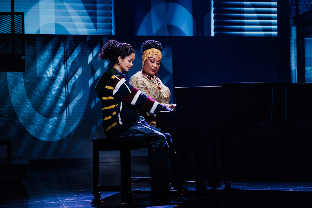 Two women performing together at a piano on stage, one musician playing and the other observing, with a colorful backdrop.