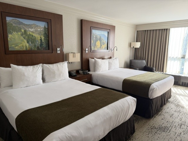 A hotel room featuring two queen beds with white linens and olive green bed runners, accompanied by two paintings above the headboards. A chair, lamp, and window with sheer curtains are visible, allowing natural light to enter the room.