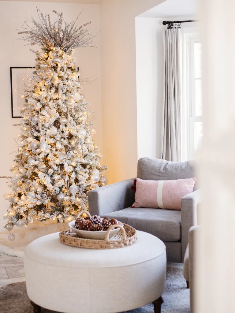 A cozy living room featuring a beautifully decorated white Christmas tree with lights and ornaments. In the foreground, there's a round coffee table with a decorative tray holding pinecones, and a grey armchair with a pink cushion.