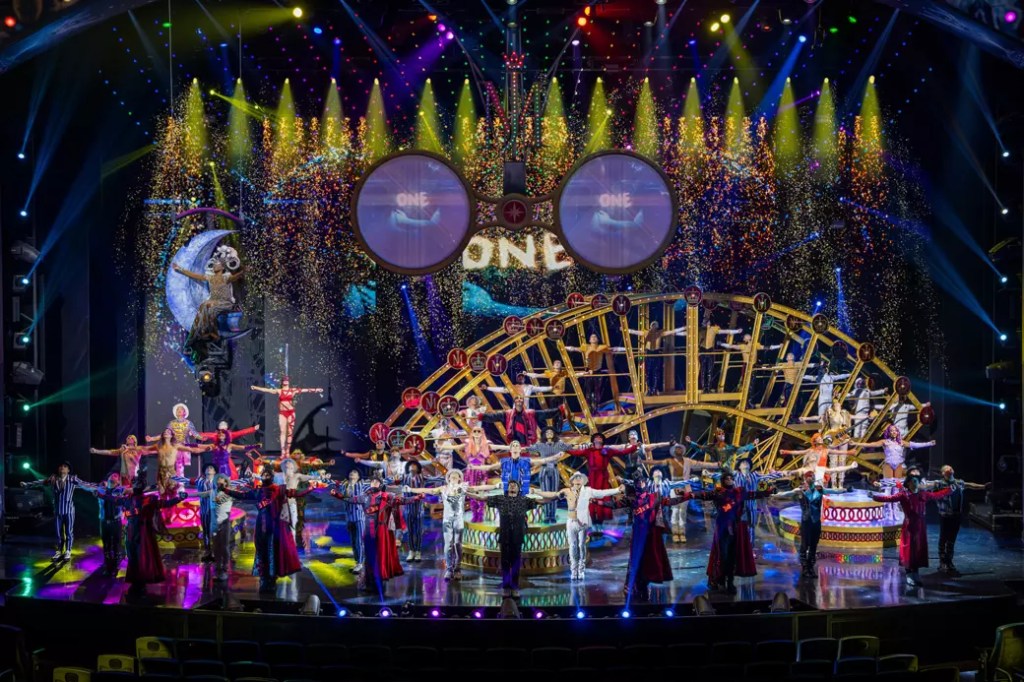 A vibrant stage performance featuring a large ensemble of performers in colorful costumes, with dramatic lighting and a backdrop of a decorative wheel and visual effects.