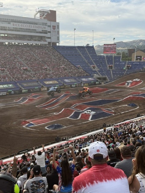 Two monster trucks performing at a motorsport event in a stadium filled with cheering spectators, with a dirt track featuring an elaborate design.