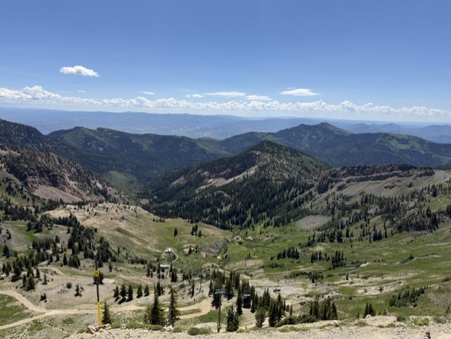 A panoramic view of a mountainous landscape featuring rolling hills, green vegetation, and a clear blue sky with scattered clouds.