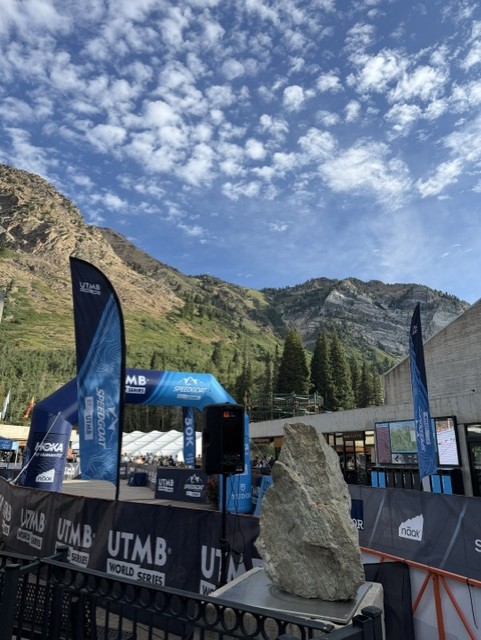 Outdoor view of a mountain landscape with a blue sky and clouds, featuring a race start/finish arch for the UTMB World Series, surrounded by banners and a large rock in the foreground.