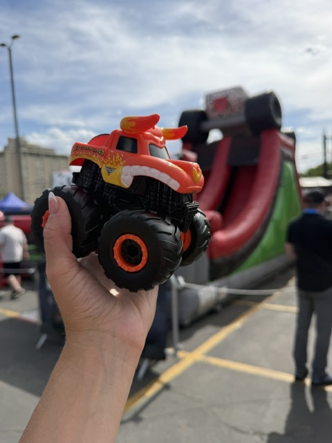 A person holding a toy monster truck in front of a large inflatable slide at an outdoor event.