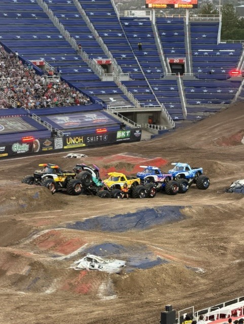 A lineup of colorful monster trucks performing on a dirt track in a stadium filled with spectators.