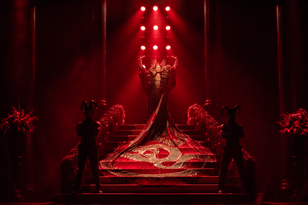 A performer in an elaborate red costume with a long train stands on an ornate staircase, flanked by two figures wearing black outfits and horned headpieces, under dramatic red lighting.