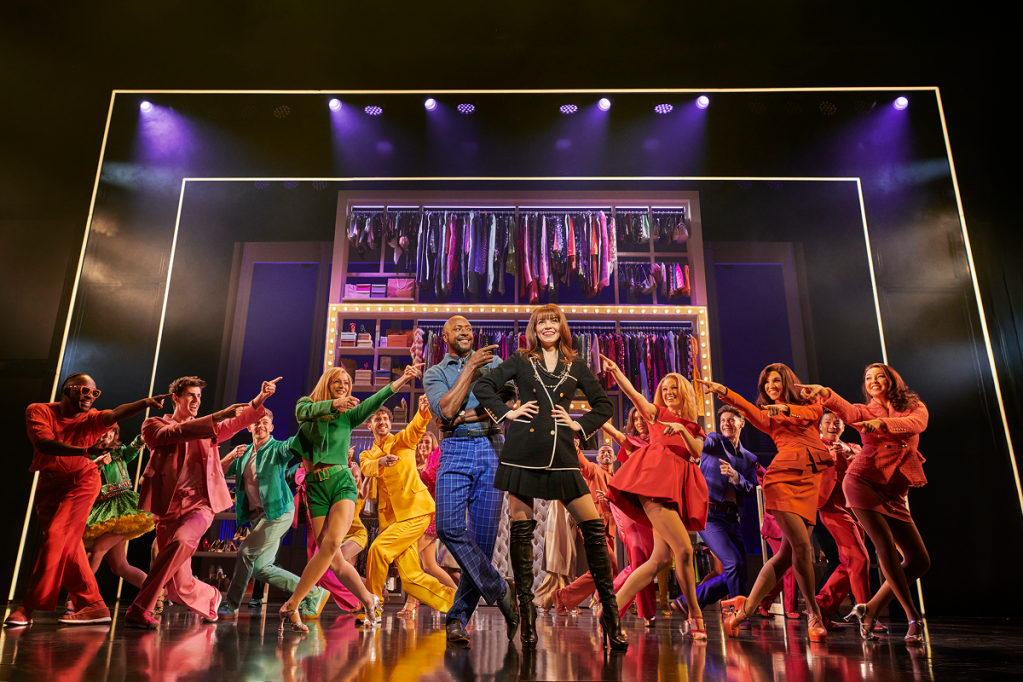 A group of performers in colorful costumes dancing on stage, with a stylish woman and a man in the foreground. A wardrobe display is visible in the background, illuminated by stage lights.