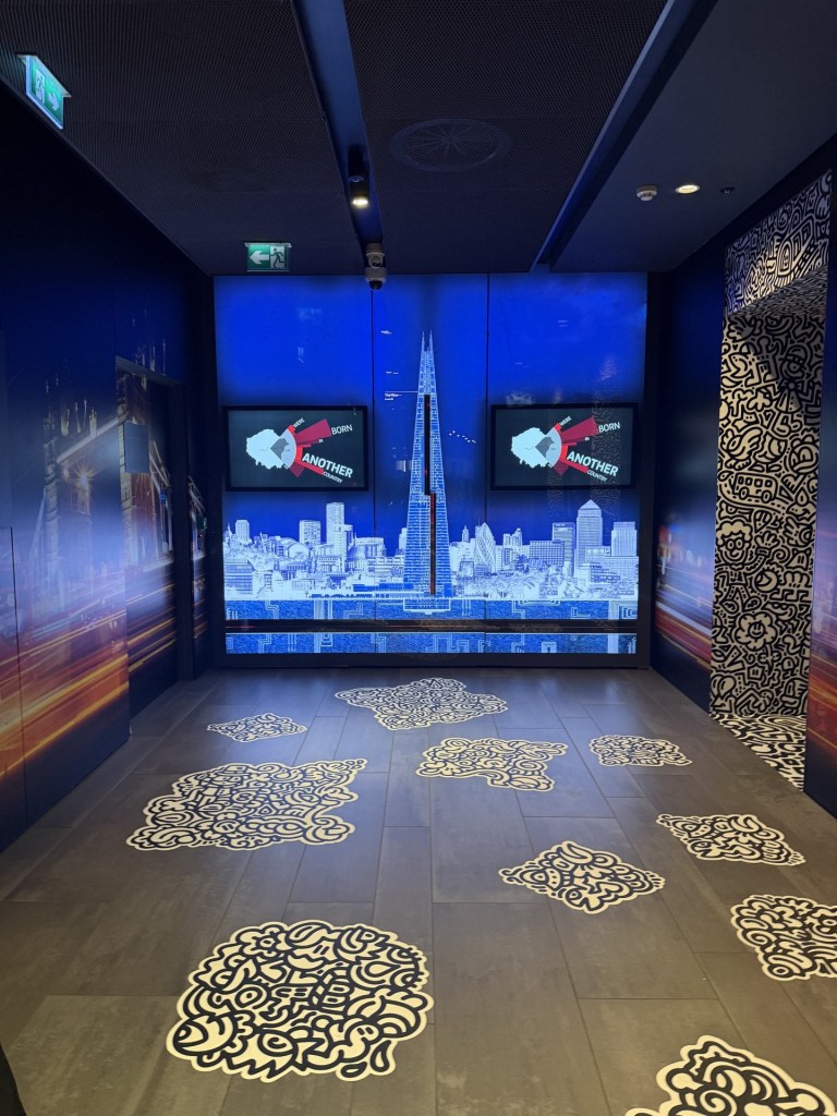 Interior view of a modern exhibition space featuring a large digital display of a city skyline, highlighted by the Shard building, against a blue background. Intricate patterned designs on the floor add artistic detail to the setting.