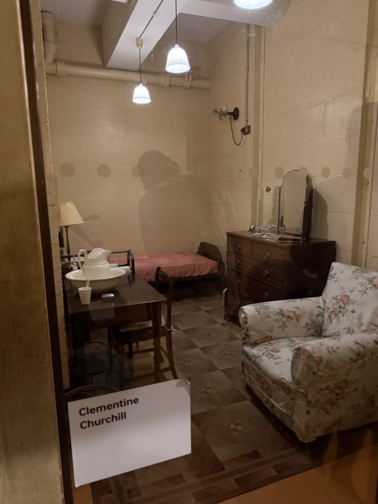 A recreated historical room belonging to Clementine Churchill, featuring a bed, a wooden table with chairs, a dresser, a floral armchair, and a lamp.