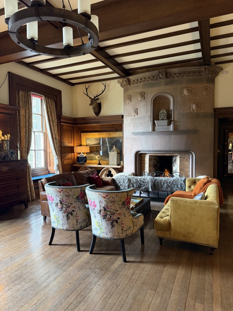 Cozy living room with a stone fireplace, floral chairs, and a plush sofa, featuring wooden beams and a deer head mount on the wall.