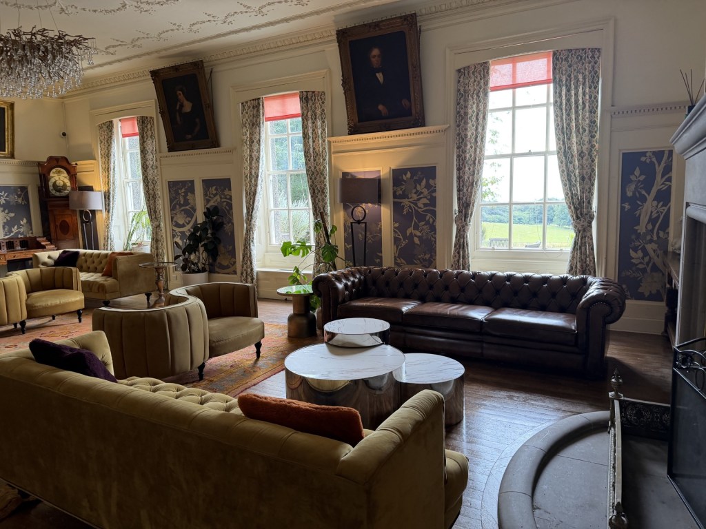 A stylish living room featuring a mix of elegant furniture, including a brown leather Chesterfield sofa, yellow upholstered chairs, and modern round tables. The room is decorated with floral wallpaper, classic portraits on the walls, and large windows with drapes, allowing natural light to fill the space.