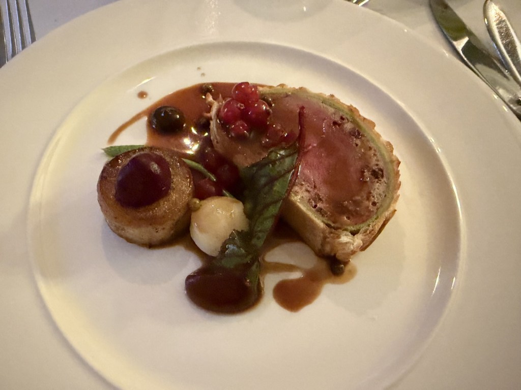 A gourmet dish featuring a slice of meat encased in pastry, garnished with a variety of colorful fruits, including berries and a green leaf, served on a white plate with a rich sauce.