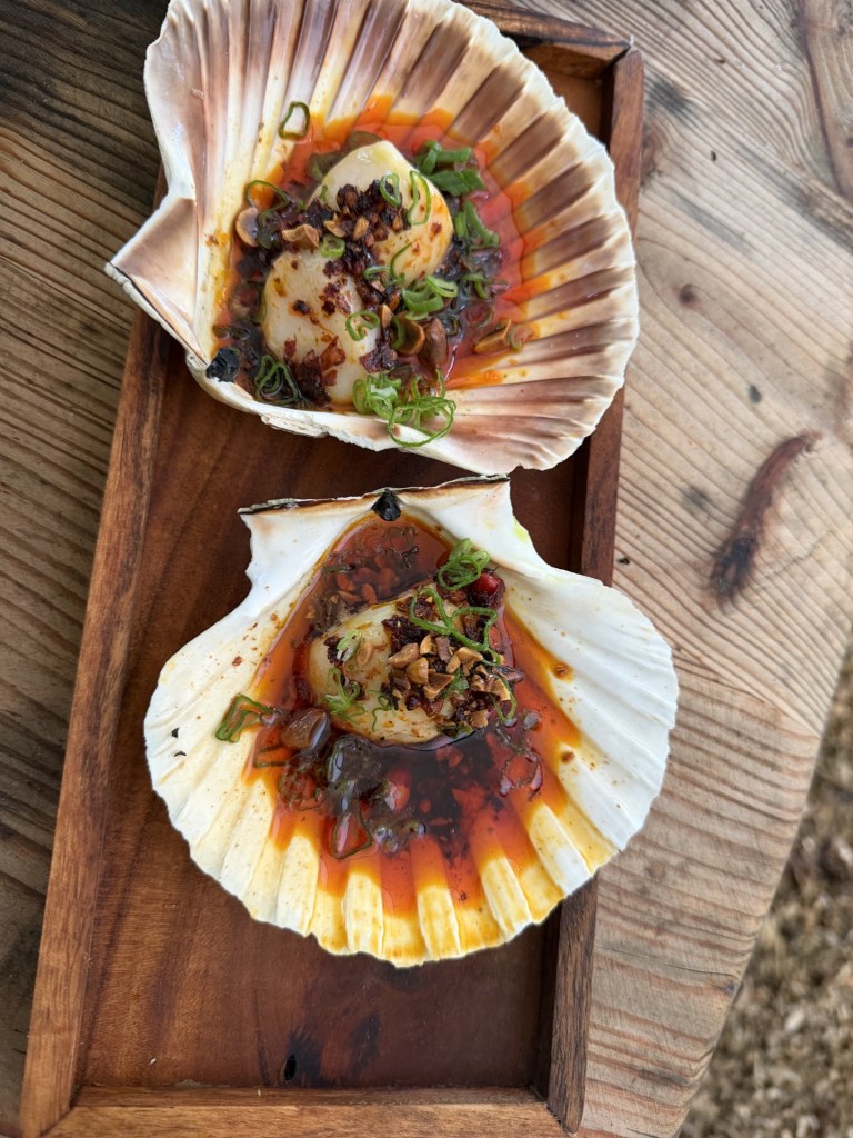 Two scallops served in their shells, topped with chili oil, minced garlic, and green onions, placed on a wooden serving tray.