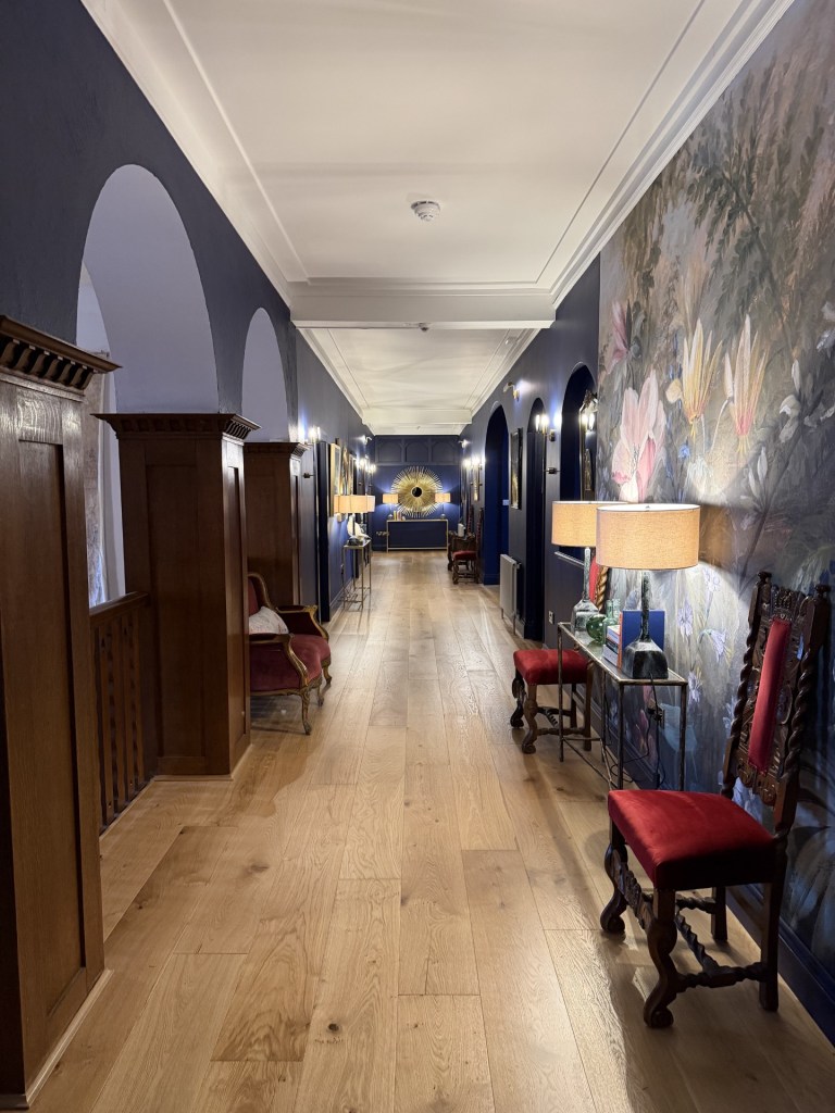 A long hallway featuring dark blue walls, wooden flooring, ornate furniture, and decorative lamps, with an art piece depicting flowers on one side.