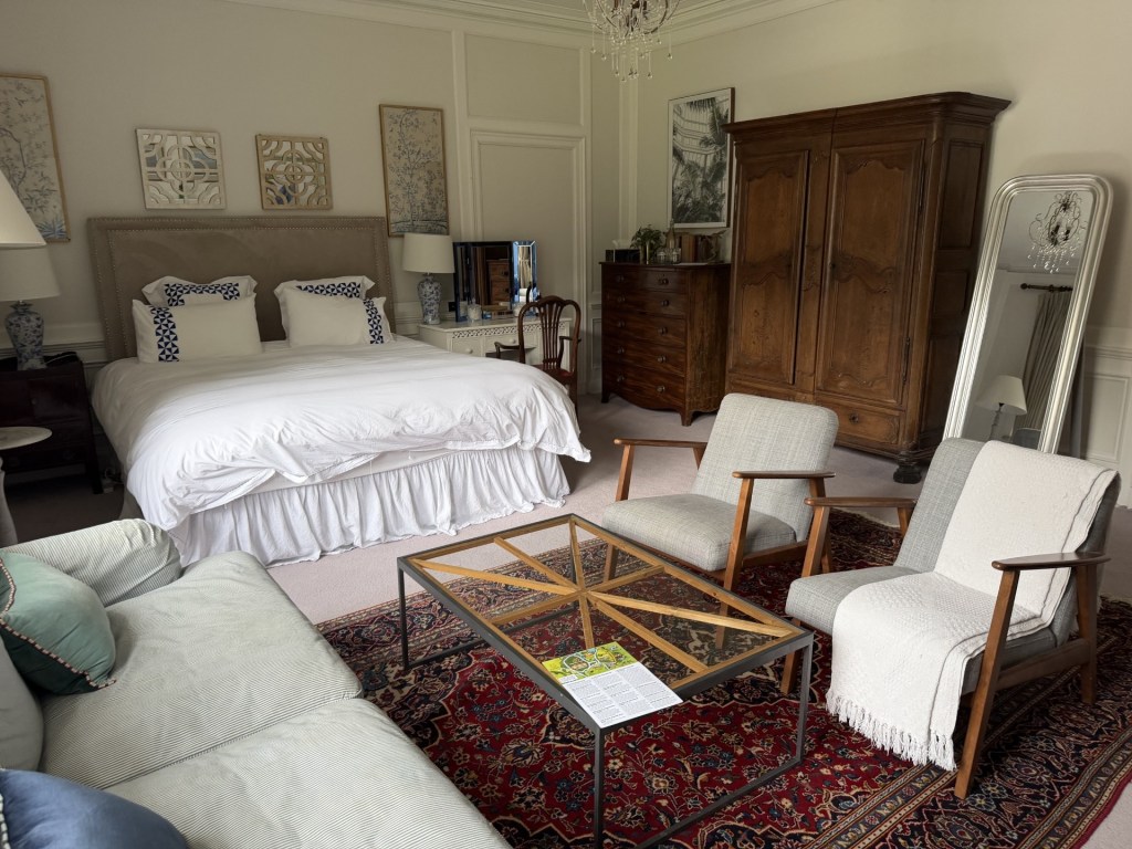 A stylish bedroom with a large bed featuring white linens and blue accent pillows, antique wooden furniture, two upholstered chairs, a glass coffee table on a patterned rug, and decorative wall art.