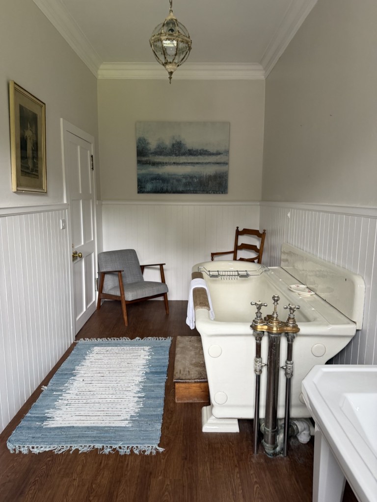 A vintage-style bathroom featuring a clawfoot tub with brass fixtures, a small wooden chair, and a gray upholstered armchair. The walls are painted in light colors with paneling, and a landscape painting hangs above the tub. A woven rug is placed on the wooden floor.