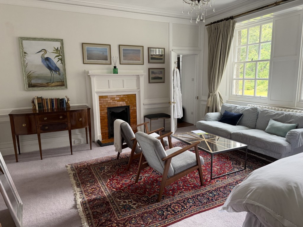 A cozy room with a vintage design featuring a patterned rug, a comfortable sofa with cushions, two armchairs, a glass coffee table, and wooden furniture. The walls are adorned with framed artwork, including a painting of a bird. A fireplace is situated in the room, with large windows allowing natural light to fill the space.
