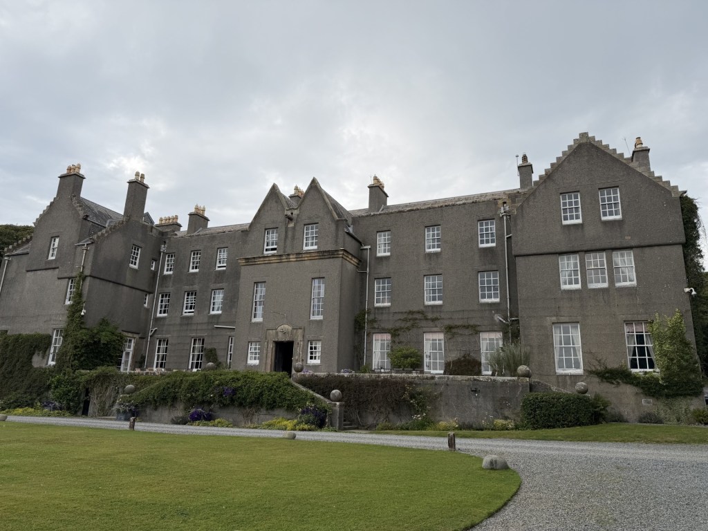 A large, historic stone mansion with multiple windows and a grassy lawn in front, set against a cloudy sky.