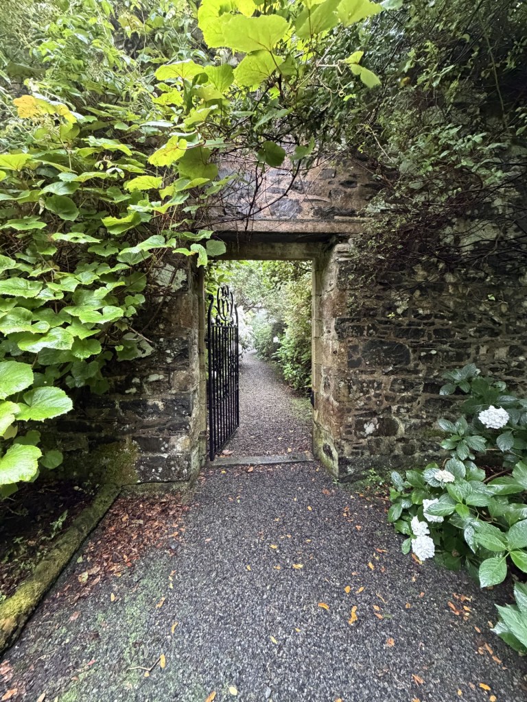 An open gate framed by lush greenery and stone walls, leading to a gravel path through a garden.