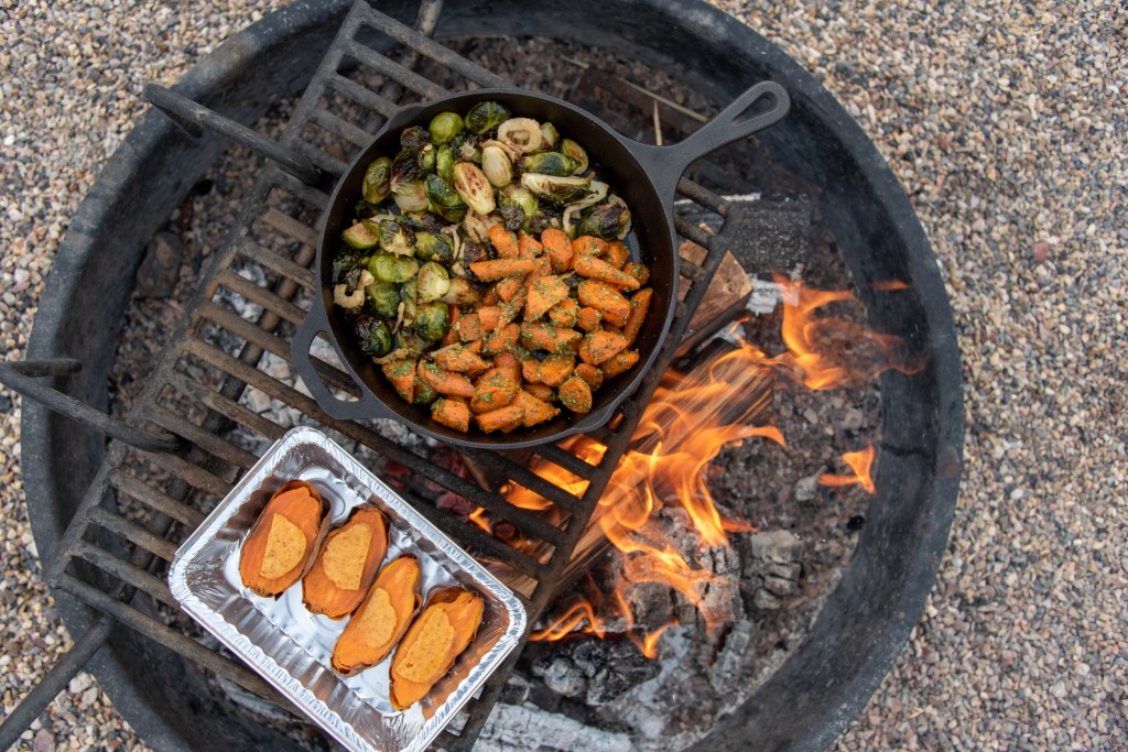 A cast iron skillet with sautéed Brussels sprouts and carrots cooking over an open fire, next to a tray of sweet potato slices.