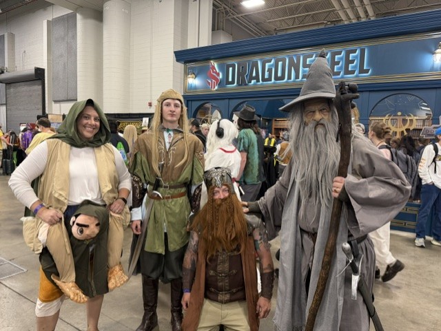 A group of four individuals in costume at a convention, featuring characters reminiscent of fantasy themes. The first person on the left is dressed in a depiction of a hobbit, the second resembles a medieval elf, the third is styled as a bearded warrior, and the fourth looks like a wizard. The background includes a vendor booth labeled 'DRAGONSTEEL'.