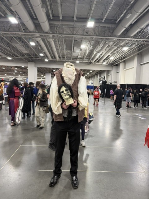 A person in a costume resembling a headless figure holding a creepy mask, surrounded by a crowd at a convention.