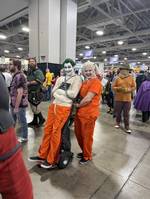 Two individuals in costume at a convention, one dressed as the Joker in orange prison attire and the other as Harley Quinn in a matching orange outfit, posing playfully together.