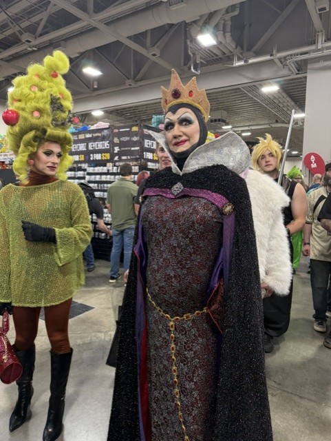 A group of individuals in colorful costumes at a convention, featuring a person in a queen costume with a crown and a dark gown, alongside another dressed in green with a unique hairstyle.