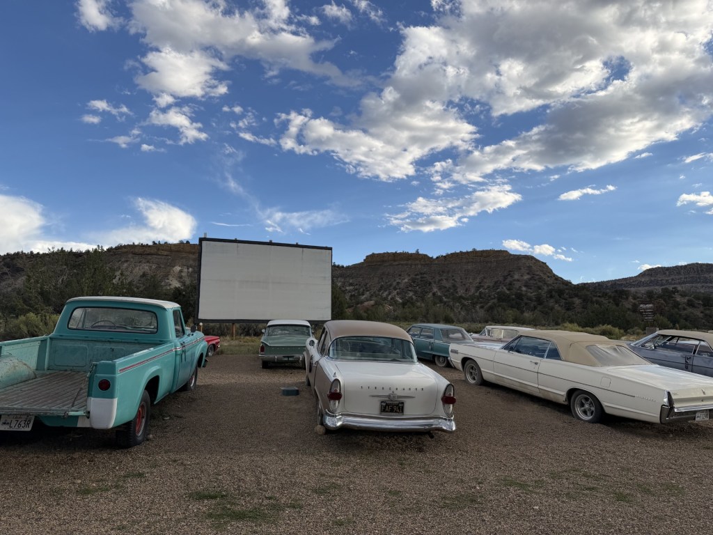 A vintage drive-in theater scene featuring several classic cars parked in front of a blank movie screen, set against a backdrop of rocky hills and a blue sky with scattered clouds.