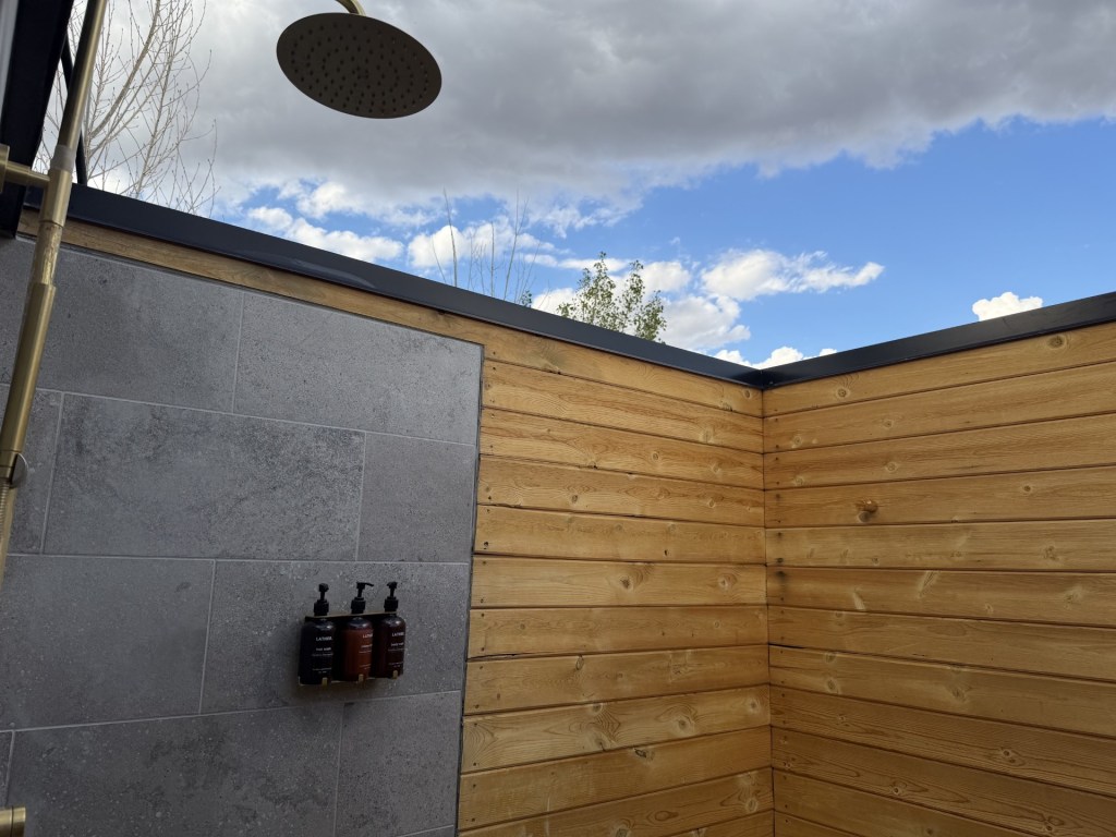 Outdoor shower with a rainfall showerhead and wooden walls, featuring gray stone tiles and three bottles of shampoo and body wash.