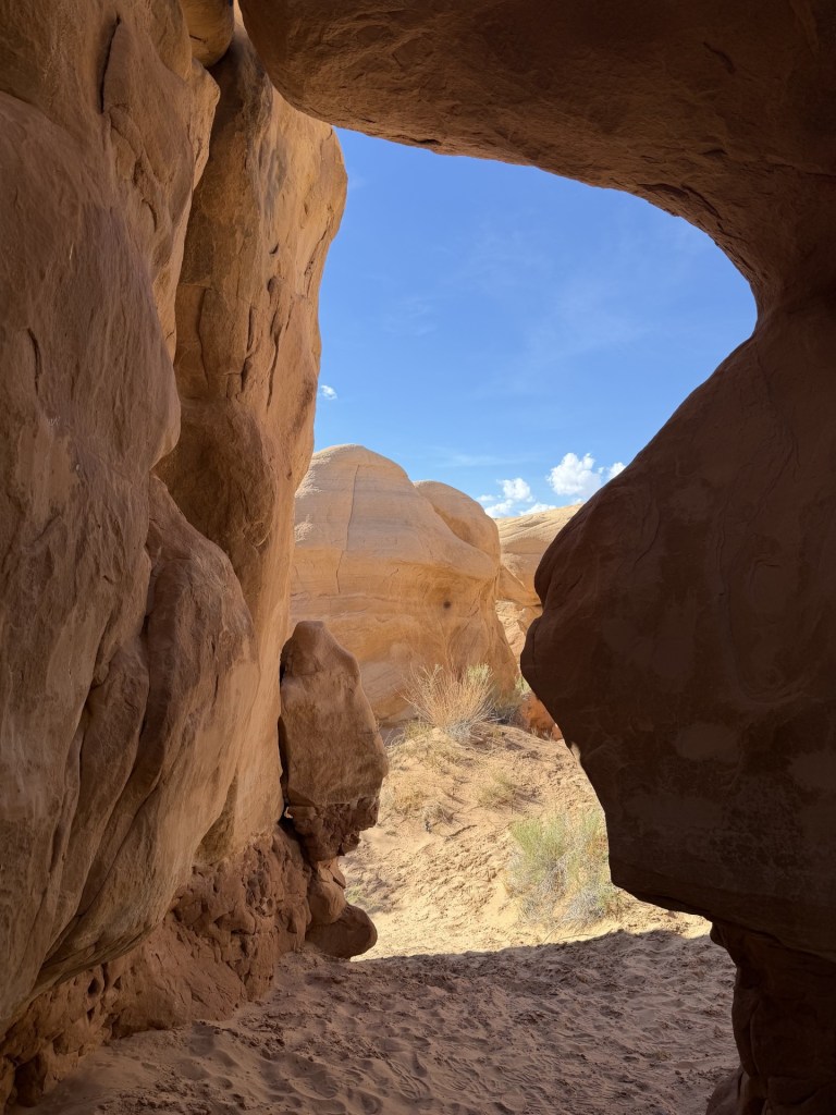 View through a narrow rock formation, revealing a clear blue sky and sandy terrain beyond.
