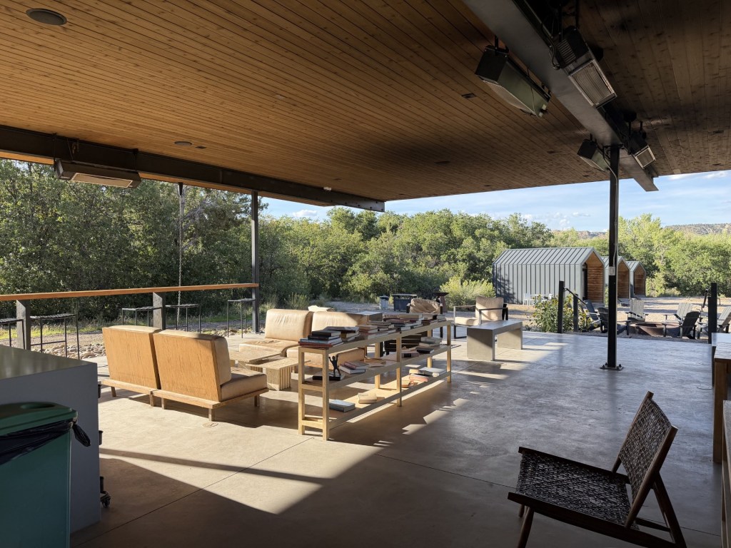 Spacious outdoor seating area with wooden furniture and tables, surrounded by greenery and a shed in the background.