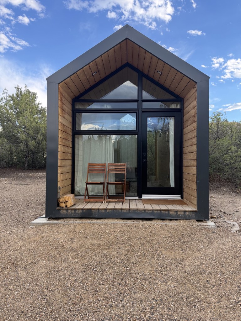 A modern small cabin with a triangular roof, featuring large glass windows and wooden accents, surrounded by gravel and sparse vegetation.