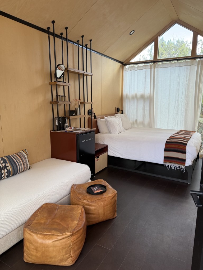 A contemporary room featuring a double bed, a seating area with a white couch, and two brown poufs. The interior has wooden walls and large windows allowing natural light. A shelving unit with a mirror and decorative items is also visible, along with a small fridge and other amenities.