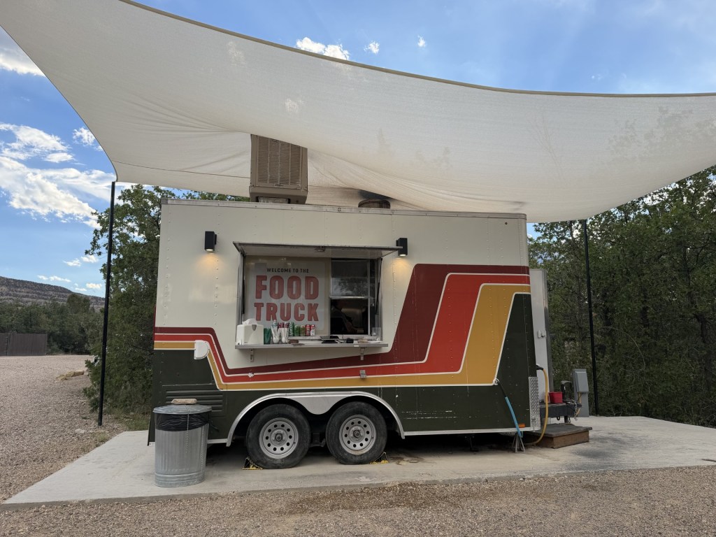 A colorful food truck with a signage reading 'WELCOME TO THE FOOD TRUCK,' located in an outdoor area under a canopy, surrounded by trees and blue sky.