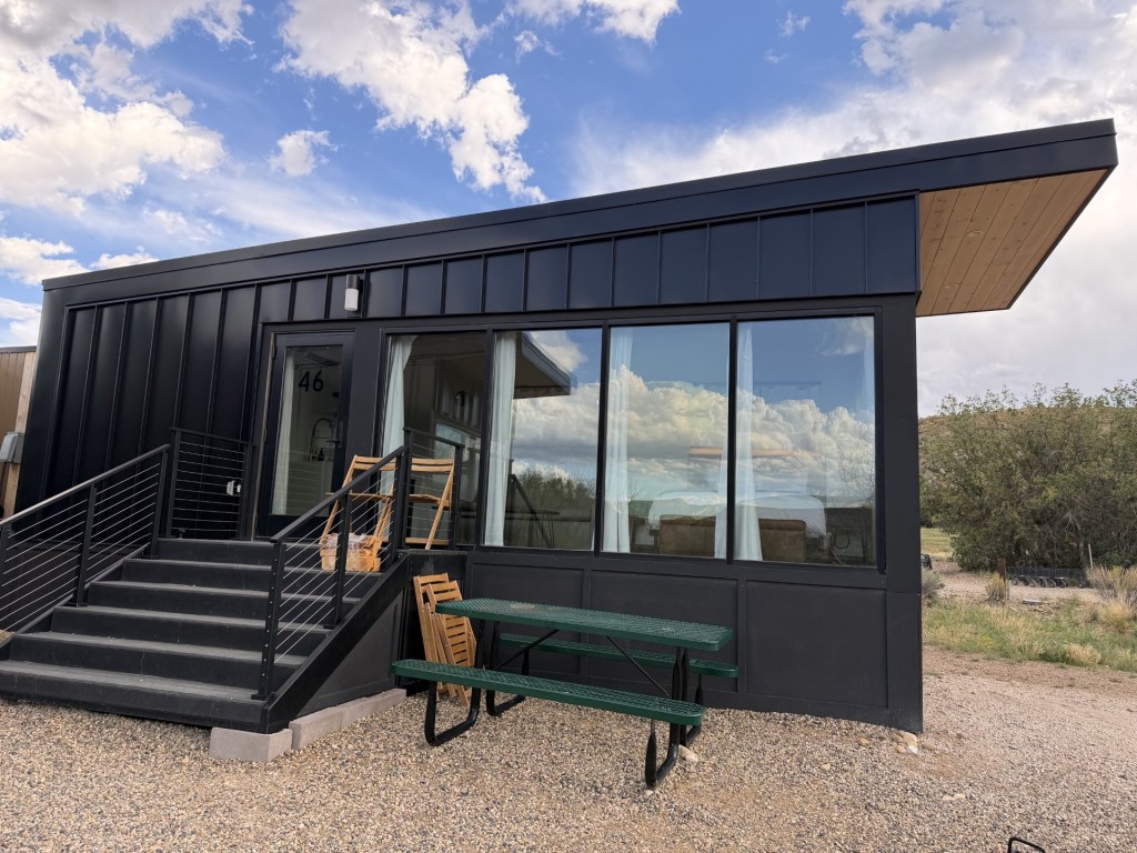 A modern black cabin with large windows, a wooden overhang, and a staircase leading to the entrance. There is a green picnic table in front, set against a backdrop of cloudy skies.