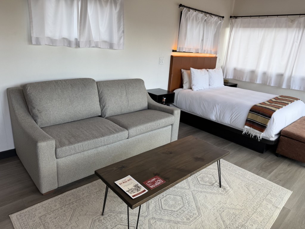 Cozy interior of a room featuring a gray sofa, wooden coffee table, and a bed with white bedding. Natural light filters through sheer curtains.