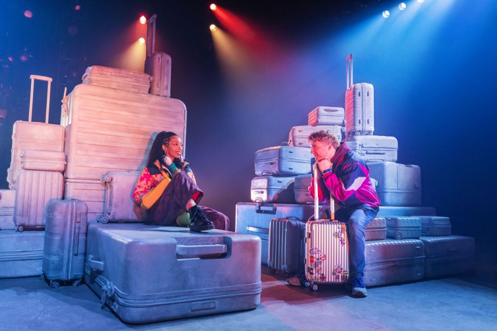 A scene from a theatrical performance featuring two actors sitting on large gray suitcases; one actor is a woman with curly hair wearing a colorful jacket, while the other is a man in a sporty jacket holding a small suitcase. The stage is illuminated with colorful lights.
