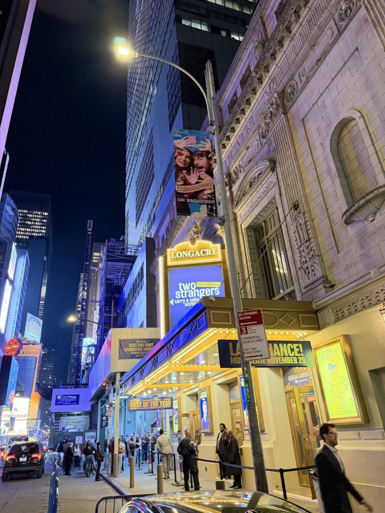 Exterior view of the Longacre Theatre at night, with illuminated marquee and promotional banners for the show 'Two Strangers Carry a Cake Across'.