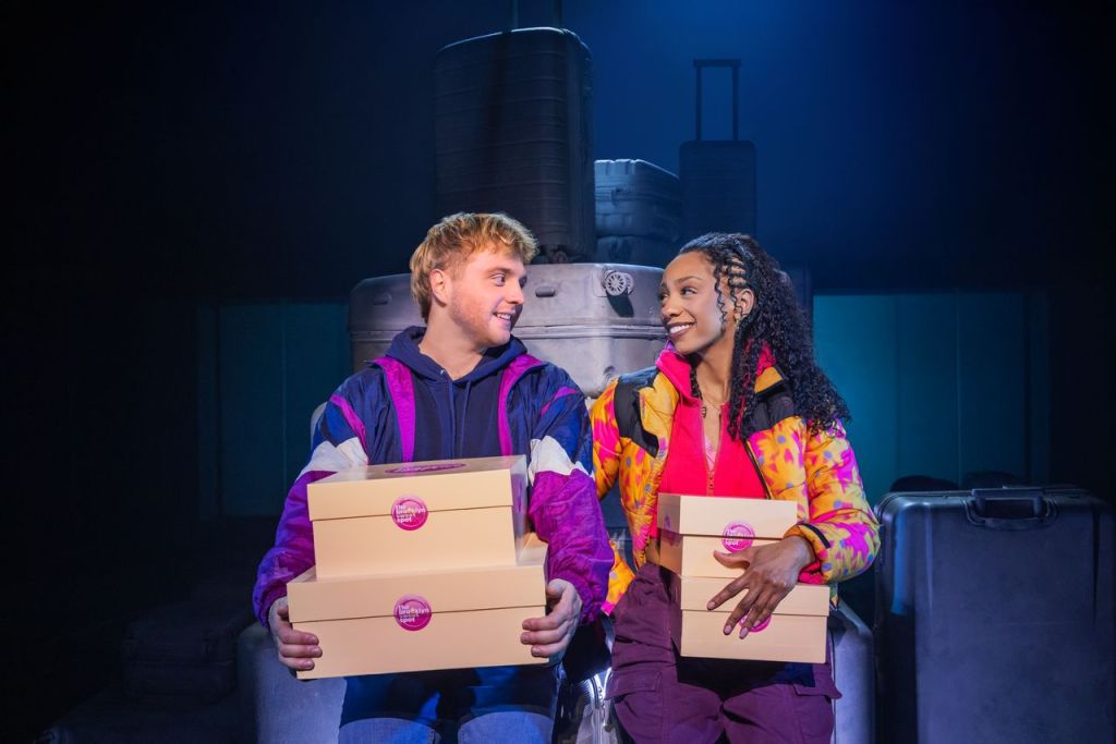 Two young adults sitting on a stack of luggage, smiling at each other while holding pink dessert boxes in a dimly lit, theatrical setting.